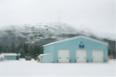 A big, baby blue shed on the side of the snowy road, with mountains in the background.