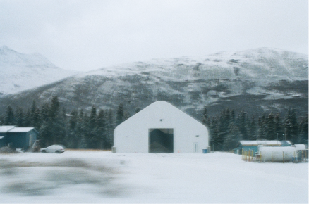 A small white shed on the side of the snowy road, with mountains in the background.