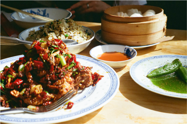 Blurry shot of a bright-green okra dish, a fried fish dish topped with red chili peppers, with a plate of fried rice and a basket of dumplings in the background.