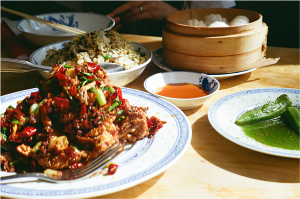 Blurry shot of a bright-green okra dish, a fried fish dish topped with red chili peppers, with a plate of fried rice, and a basket of dumplings in the background.