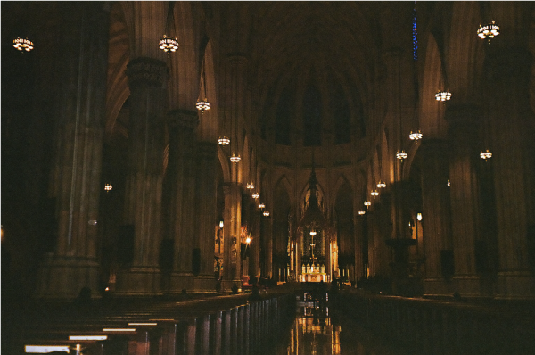 Dark shot of the interior of a cathedral, the pews and the altar lit by candlelight.