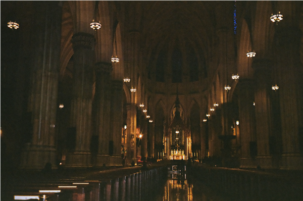 Dark shot of the interior of a cathedral, the pews, and the altar lit by candlelight.