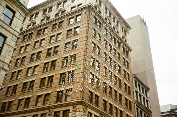 Art installation of iridescent geometric shapes together form a mesh wire, hung amidst the buildings.