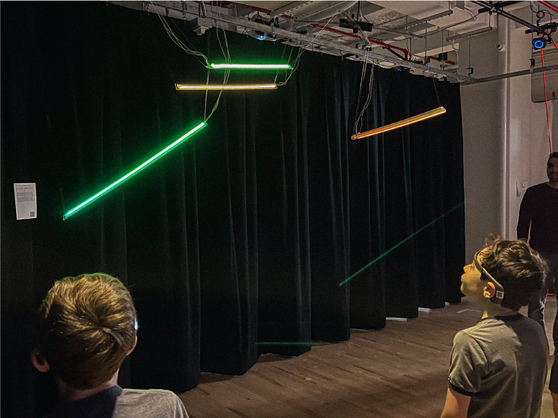 Two kids with black EEG headsets on stand in front of 2 pairs of LEG lights suspended from the ceiling--the pair of lights on the left shine green and the two on the right shine orange.
