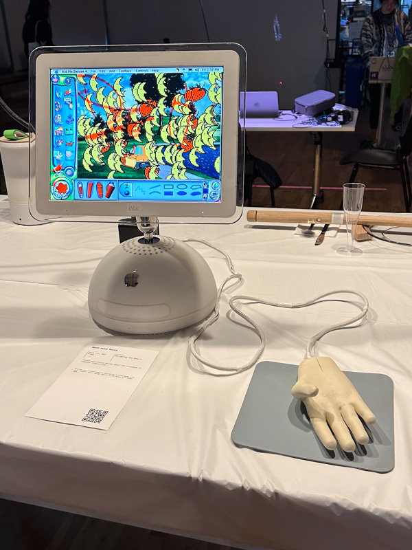 A table display with the handhold mouse, on top of a light blue mousepad, connected to a iMac G4 computer--people to play with an old drawing program called Kid Pix (2000) on the iMac.