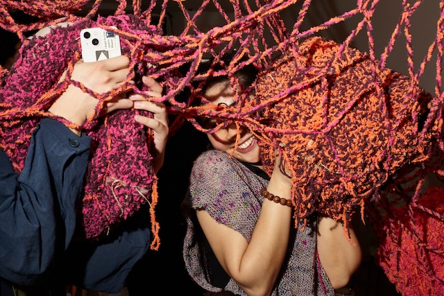 A pink, orange, and dark purple net cascades from the ceiling, obscuring two people hugging nodes inside the net, one person is holding a phone, and the other person is smiling.