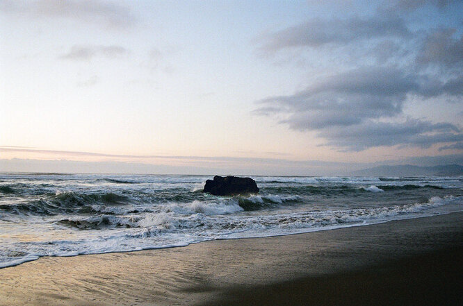 Waves crashing onto a lone rock on an empty beach, somewhat overcast clouds in the sky.